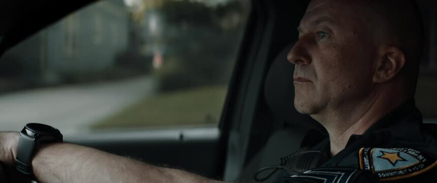 CU Caucasian American police officer patrolling streets of neighborhood in patrol vehicle. Shot with 2x anamorphic lens