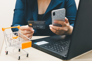 A woman holding a credit card and smartphone, which is paying online, sits on the chair in the living room at home on a laptop. The concept of online shopping and e-commerce..