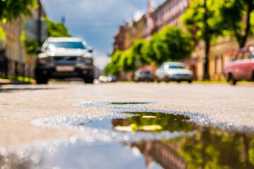 Sunny day after the rain in the city, parked on the street cars on the background of the facades of the house and trees. Close up view from the level of the puddle on the pavement
