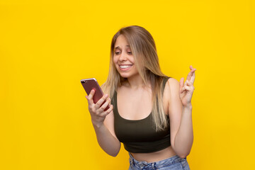 Naklejka premium A young pretty caucasian smiling blonde woman crosses her fingers for good luck looking at the phone screen waiting for the results of the lottery or exams isolated on a bright color yellow background
