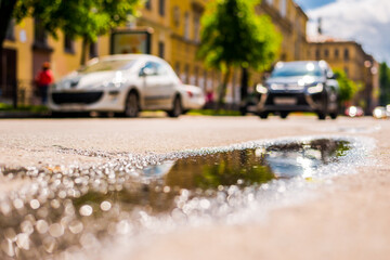 Sunny day after rain in the city, the car rides along the road. Close up view from the level of the puddle on the pavement