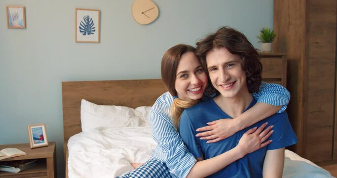 Love and tenderness. Image of optimistic smiling young loving couple sitting at the bed indoors at home and hugging. Husband and wife enjoying of free time with each other. Stock video