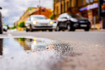 Sunny day after rain in the city, parked cars near the store and a passing car. Close up view from the level of the puddle on the pavement