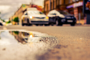 Sunny day after rain in the city, parked cars near the store and a passing car. Close up view from the level of the puddle on the pavement