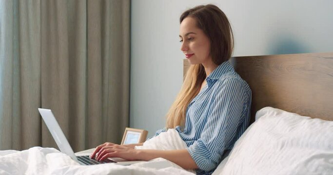 Side view of the busy woman sitting at the bed and typing at the laptop keyboard while working remotely from home during the coronavirus pandemic. Pleasant woman using computer at the bed