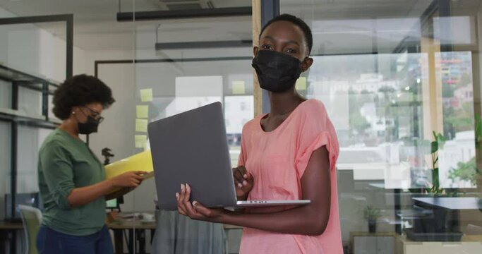 Happy african american businesswoman wearing face mask using laptop, looking to camera in office