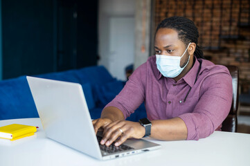 Concentrated African-American male employee wearing medical mask sitting at the desk and typing, working with laptop computer in home office, protective measures and social distancing during pandemic