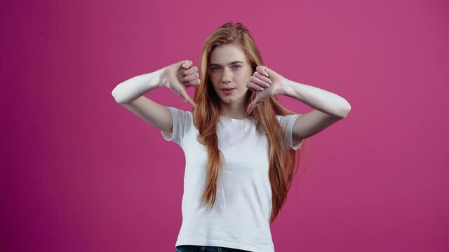 The portrait of a dissatisfied redhead who shows the sign of dislike with both hands. Freckled teenage girl in a white T-shirt, isolated on a pink background. The concept of people's lifestyle