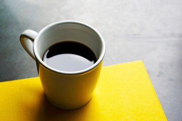 A cup of black coffee on yellow paper and black table background with window light.