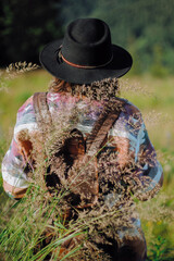 Woman traveler with backpack and hat gathering wildflowers in grass on mountain hills, back view