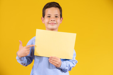 Smiling boy portrait holding white blank paper. Isolated on yellow background