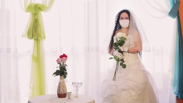 A Jewish bride in a wedding dress and a veil in a medical mask in the synagogue hall with a bouquet of white roses turns her face.