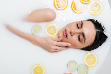 top view of young woman touching face while bathing in milk with citrus slices.