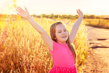 A little smiling girl with long hair in a pink dress stay along a country road. Happy childhood, lifestyle, cozy mood, summer vacation. concept