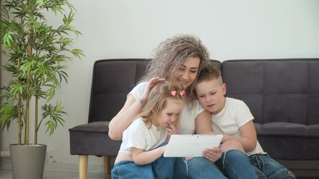 Mom and daughter and son play with the tablet in a cozy living room. The family is dressed in the same basic white T-shirts. Leisure with children. Family entertainment