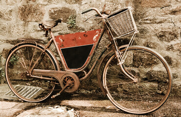 Obraz na płótnie Canvas Rusty vintage bicycle with black board for entering a text (advertisement, menu etc) and wicker basket leaning on a stone wall. Brittany, France. Sepia toned photo.