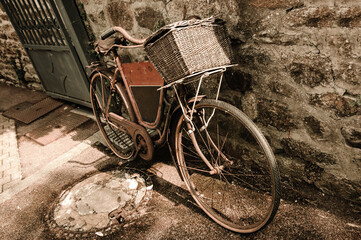 Fototapeta na wymiar Rusty vintage bicycle with black board for entering a text (advertisement, menu etc) and wicker basket leaning on a stone wall. Brittany, France. Sepia toned photo.