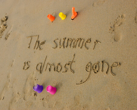A sentence written on the sand of a beach: THE SUMMER IS ALMOST GONE,  four colorful molds and shovel . Vacation end / back from holiday / back to the work or school concept.