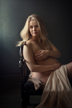 Beautiful pregnant woman wrapped in piece of fabric with naked belly sitting on the bed in dark room. Image with selective focus, toning and noise effect.