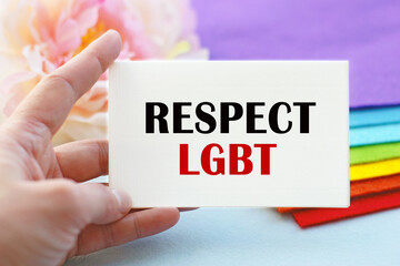 A woman's hand holds a piece of paper with the inscription RESPECT LGBT, against the background of a stack of cloth folded in the colors of the LGBT flag. LGBT concept.