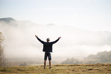 Happy traveler man raising hands up on the top of the world above mountains in white clouds. Hipster guy enjoy amazing atmospheric moment. travel and wanderlust concept