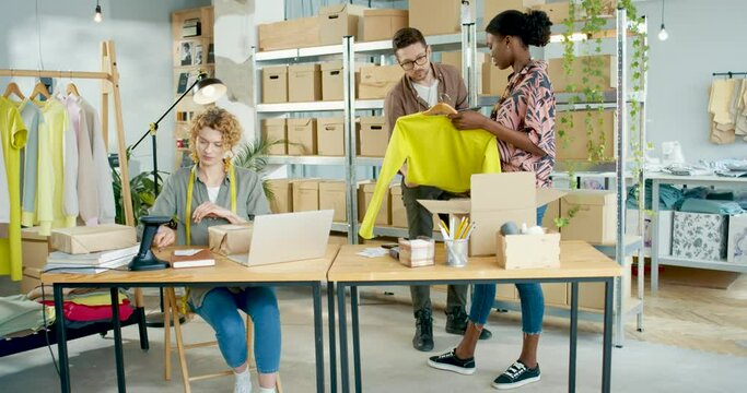 Stylish mixed-race young people working in fashion industry in clothing store. Male manager tapping on tablet, female stylist designer packing clothes in warehouse. Woman scanning parcel using scanner
