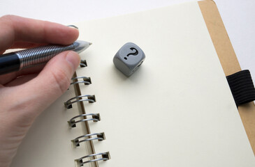 Left hand holding a pen above a blank page of a spiral bound notebook, gray dice with question mark...