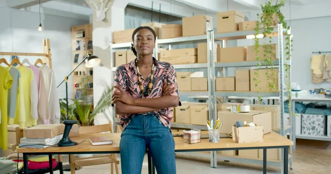 Cheerful positive African American young beautiful businesswoman with dreamy face standing in office in own small clothing store and looking at camera smiling in warehouse, stylist, business concept