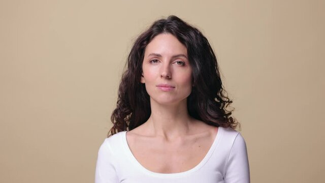Head shot of happy smiling beautiful millennial woman in white casual clothes over beige background. Young adult female with curly brunette hair and toothy smile. Lifestyle, sincere emotion concept