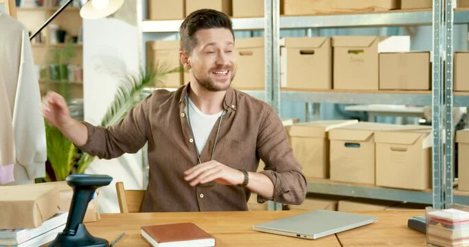 Portrait of happy young handsome man sitting at desk at office in clothing shop warehouse speaking on video call looking at camera. Caucasian smiling male stylist recording video blog