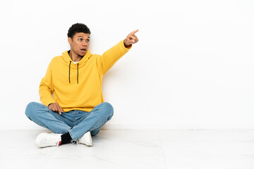 Young African American man sitting on the floor isolated on white background pointing away