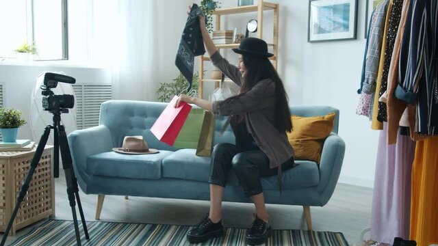 Asian stylist blogger is taking scarf from shopping bag and wearing it recoding video for social media talking and smiling using camera on tripod.