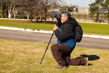 An adult man, crouching with a camera mounted on a monopod, in the park with green areas.