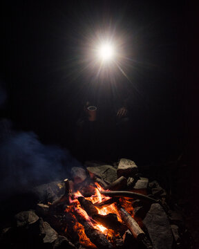 A headlamp shines from a person around a campfire at night