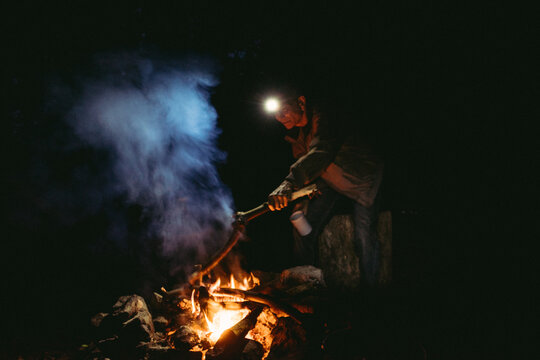 An older man 55-70 years old places a log on a campfire at night