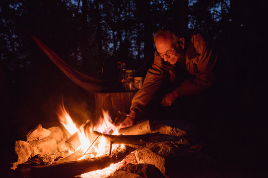 An older man 55-70 years old places a log on a campfire at night