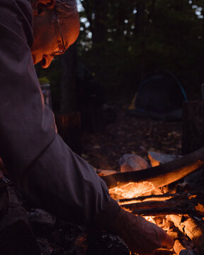An older man 55-70 years old places a log on a campfire at night