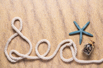 Fototapeta na wymiar Sea spelled out with nautical rope with a blue starfish and small glass bottle of seashells on a beach sand background