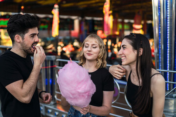 Dos chicas y un chico amigos sonriendo y compartiendo un algodón de azúcar rosa en zona de atracciones de una feria