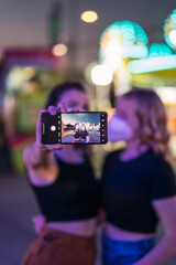Fototapeta na wymiar Dos chicas rubia y morena tomando selfies en zona de atracciones de una feria