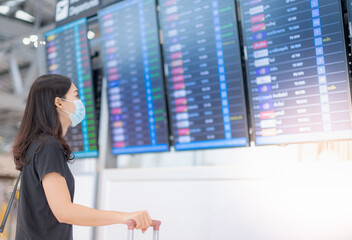 Asian woman wearing surgical mask and holding passport in her hand and luggage looking at the flight information board, checking her flight at the airport.