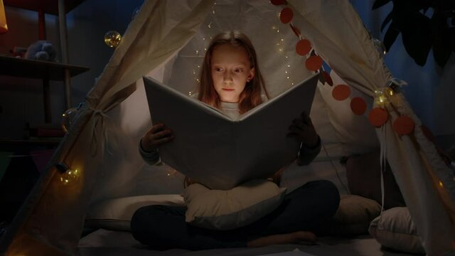 Amazed girl with red hair in makeshift decorative tent opening book at home in evening. Teen making surprised face while sitting on floor and reading book with flashlight.