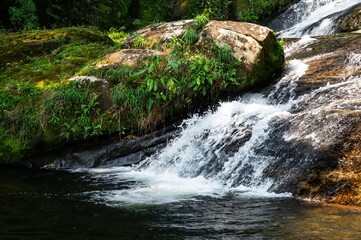 Naklejka premium Water current flowing around rock formations covered in moss of Ipiranguinha waterfall forming a golden natural pool right below in Serra do Mar (Sea Ridge) forest in Cunha, Sao Paulo - Brazil.