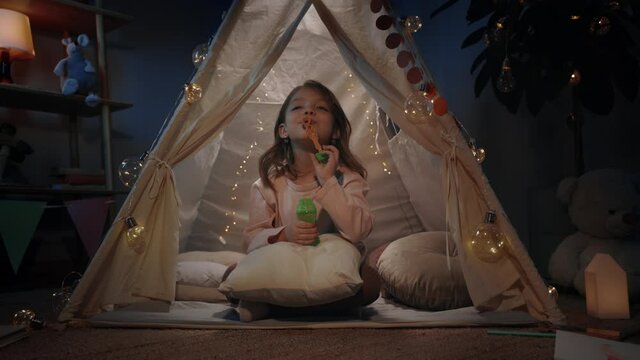 Adorable kid girl in beautiful decorative tent blowing bubbless and looking to camera in evening. Little child sitting on floor while having fun in her room. Concept of childhood.