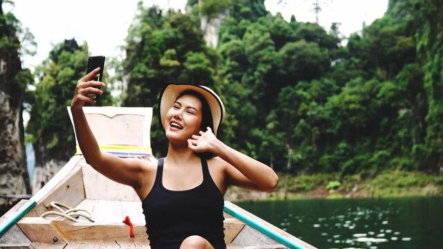 Travel adventure, Freedom, experience, sightseeing concept. Cheerful young woman wear hat sitting on the wood long tail boat in tropical using smartphone selfie taking photo over nature background.