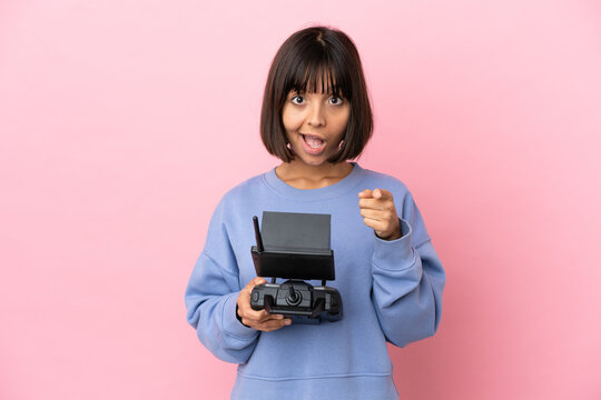 Young mixed race woman holding a drone remote control isolated on pink background surprised and pointing front