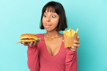 Young brunette woman holding burger and fried chips over isolated blue background