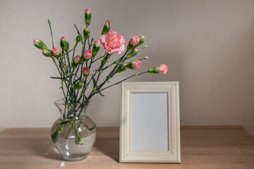 Portrait white picture frame mockup on wooden table. Modern ceramic vase with carnation. White wall background. Scandinavian interior. 