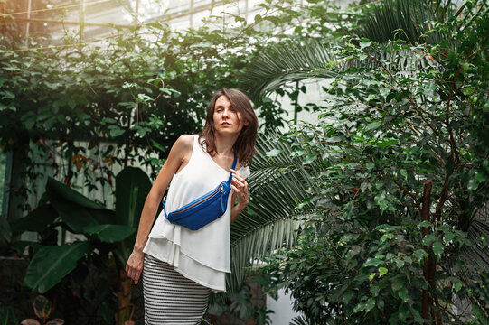 Beautiful caucasian young woman in a white blouse, skirt and blue banana bag on a background of green plants.