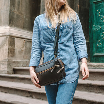 Leather sling bag for women. Belt Bag Minimalist. Blond woman in denim shirt and, blue jeans and a dark brown leather bag over the shoulder. Close-up.